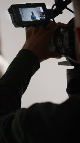 Vertical-Video-Close-Up-Of-Film-Camera-Operator-Shooting-Actors-Rehearsing-Movie-Or-Video-In-Studio-Viewed-Through-Monitor